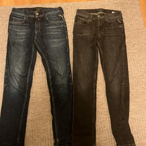 Replay jeans i blått och svart, 10A - Två par Replay jeans, ett par i mörkblå tvätt och ett par i svart tvätt. Båda har klassisk femficksdesign, raka ben och snygga detaljer på bakfickorna. Perfekta för dig som gillar stilrena och bekväma jeans med modern passform.