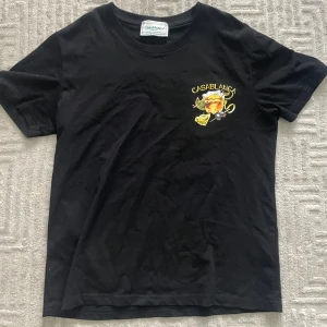 Svart Casablanca t-shirt med print - Svart t-shirt från Casablanca med färgglatt tryck av drink, citron och oliver både fram och bak. Klassisk rund hals och korta ärmar. Skön bomullskvalitet och snygg streetstil med cool grafik och gul text. Priset är inte hugget i sten! Vid fler frågor eller funderingar tveka inte på att skriva!