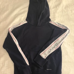 Mörkblå hoodie från GANT med logga - Snygg mörkblå hoodie från GANT med huva och vita ränder längs ärmarna där GANT-loggan syns. Röd detalj på ärmarna och insidan av kragen. Mjuk bomullskänsla och klassisk känguruficka framtill. Barn storlek.