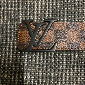 Louis Vuitton rutigt brunt skärp - Säljer ett Louis Vuitton bälte i klassiskt brunt och mörkbrunt rutigt mönster med stor LV-logga i svart metall framtill. Bältet är gjort i äkta skinn och har exklusiv känsla, perfekt för dig som vill sticka ut med en ikonisk accessoar.