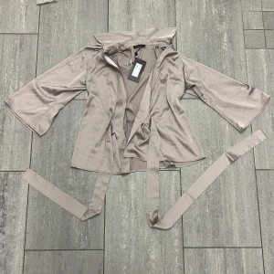 Silvrig omlottjacka från STOCKH LM - Snygg silvrig omlottjacka från STOCKH LM i modellen Axelia. Jackan har vida trekvartsärmar, bred krage och knytskärp i midjan. Tillverkad i ett glansigt, mjukt material som ger en lyxig känsla. Perfekt för dig som vill ha en stilren och trendig look.