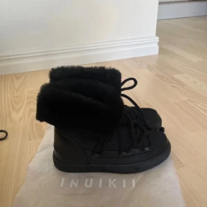Svarta Inuikii boots med päls - Säljer ett par svarta Inuikii boots med fluffig päls runt skaftet och grova snören. Skorna har en platt sula och är tillverkade i skinn och päls, perfekta för kalla dagar. Snygg och chunky design som sticker ut.