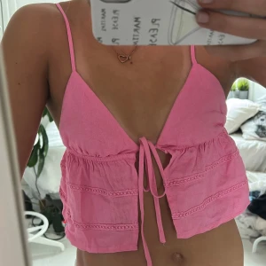 Rosa linnetopp med knyt framtill - Säljer en söt rosa linnetopp med smala axelband och djup v-ringning. Toppen är croppad och har knyt framtill samt dekorativa spetsdetaljer i horisontella rader. Perfekt för varma dagar och ger en chill vibe.