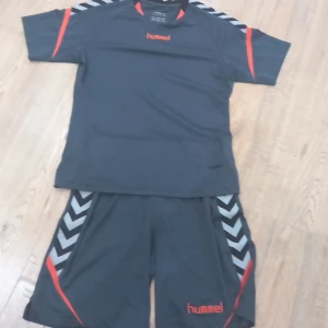 Mörkgrå sport-tshirt och shorts från Hummel - Sportig mörkgrå t-shirt och shorts från Hummel med korta ärmar, orange logga och detaljer samt svarta och grå chevronränder på axlarna. Tillverkad i lätt och ventilerande polyester, perfekt för träning eller fotboll. Snygg och funktionell design för dig som gillar sportiga vibes.