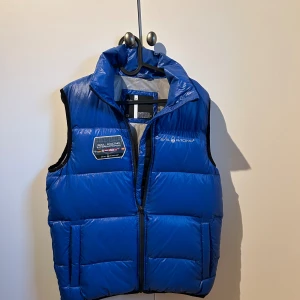 Blå dunväst från Sail Racing - Blå dunväst från Sail Racing med hög krage och dragkedja framtill. Västen har en patch med logga på bröstet och två fickor med dragkedja. Materialet är glansigt och lätt, perfekt för lager-på-lager. Snygg sportig look med tydliga sömmar och diskreta svarta detaljer.