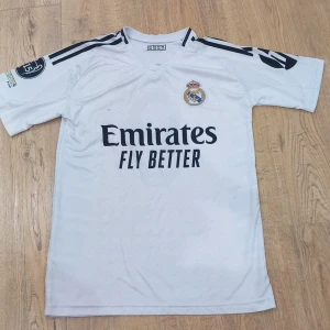 Real Madrid Mbappé fotbollströja 164 - Vit Real Madrid fotbollströja med Mbappé och nummer 9 på ryggen. Tröjan har svarta detaljer, klubbmärke på bröstet och sponsortryck framtill. Korta ärmar och tillverkad i lätt, ventilerande material. Perfekt för dig som älskar fotboll och vill sticka ut på planen.