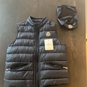 Svart dunväst från Moncler - Snygg svart dunväst från Moncler med quiltad design och hög krage. Västen har dragkedja framtill, två fickor med dragkedja och klassisk Moncler-logga på bröstet. Tillhörande påse för smidig förvaring ingår. Perfekt för lager-på-lager-stil under kyliga dagar.