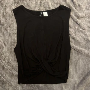 Svart twistad linnetopp från Divided - Svart ärmlös linnetopp från Divided med snygg twistad detalj framtill. Toppen har rund halsringning och är tillverkad i mjuk bomull, perfekt för en stilren och enkel look.