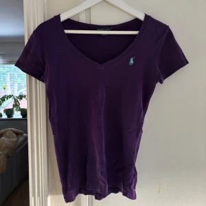 Lila t-shirt från Ralph Lauren - Snygg lila t-shirt från Ralph Lauren med v-ringning och korta ärmar. Klassisk logga broderad på bröstet i ljusblått. Perfekt till jeans eller shorts för en chill och stilren look.