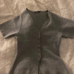 Grå stickad topp från Gina Tricot - Säljer en grå stickad topp från Gina Tricot med v-ringning och korta ärmar. Toppen har en rad med knappar framtill och är figurnära i modellen. Nyköpt, aldrig använd, inga defekter 