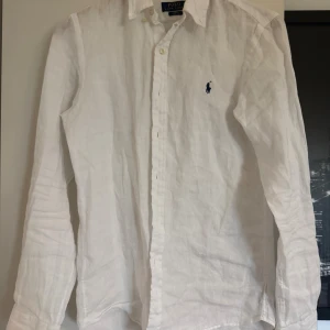 Vit linneskjorta från Polo Ralph Lauren - Klassisk vit skjorta i 100% linne från Polo Ralph Lauren. Skjortan har lång ärm, knappar framtill och den ikoniska blå broderade loggan på bröstet. Perfekt för en clean och fräsch look med avslappnad passform.