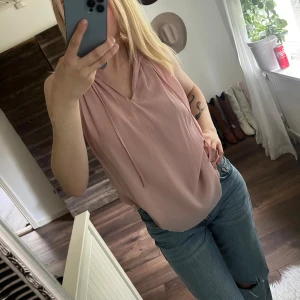 Ljusrosa ärmlös blus med v-ringning - Säljer en ljusrosa blus med v-ringning och tunna knytband vid halsen. Blusen är ärmlös och har en lös passform, perfekt till jeans eller kjol. Materialet känns lätt och luftigt, vilket gör den skön att bära under varmare dagar.