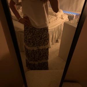 Blommig långkjol med spetsdetaljer - Supersöt långkjol med blommigt mönster i brunt, blått och vitt. Kjolen har flera breda spetsband som ger en bohemisk vibe och är perfekt för dig som gillar romantiska detaljer. Flödande passform och mjukt tyg som känns skönt mot huden.