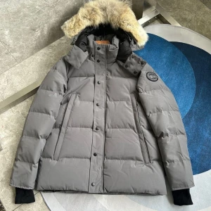 Grå dunjacka med fuskpäls från Canada Goose - Säljer en grå dunjacka från Canada Goose med stor huva och fuskpäls. Jackan har svarta muddar, raka fickor och patch-logga på ärmen. Perfekt för kalla dagar och har en klassisk, stilren look.