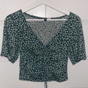 Grön blommig croppad blus H&M - Söt grön blus med vitt blommönster från H&M. Toppen har v-ringning, korta ärmar och en croppad passform med snörning framtill. Perfekt för vår och sommar med sitt lätta material och trendiga look.