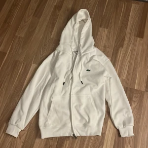 Vit Lacoste Hoodie - Vit hoodie från Lacoste. Tröjan har hel dragkedja, snörning i huvan och två fickor framtill. Tillverkad i mjukt material.