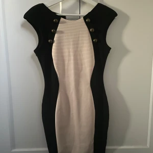Svart och beige kortklänning med knappar - Snygg kortklänning i svart och beige med markerade axlar och dekorativa knappar framtill. Klänningen har en figurnära passform och är ärmlös och stretchig i materialet! 