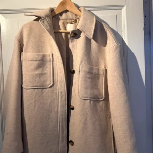 Beige overshirt med stora fickor - Stilren beige overshirt med två stora bröstfickor och knäppning framtill. Jackan har klassisk krage och raka ärmar, perfekt för lager-på-lager. Materialet känns mjukt och har en lätt struktur som ger en avslappnad vibe.