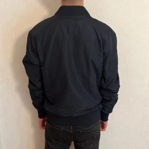 Svart bomberjacka från J.Lindeberg - Säljer en marin blå bomberjacka från J.Lindeberg i storlek M. Jackan har klassisk passform, ribbade muddar och silverfärgad dragkedja framtill. Tillverkad i ett slätt syntetmaterial som känns lätt och smidigt. Perfekt för dig som gillar stilren och enkel design.  