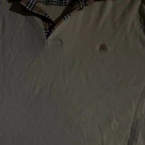 Beige pikétröja från Burberry - Snygg beige pikétröja från Burberry med klassisk rutigt mönster på insidan av kragen och broderad logga på bröstet. Tröjan har en stilren design och är tillverkad i mjuk bomull. Perfekt för dig som gillar preppy och tidlös stil. Passar som M