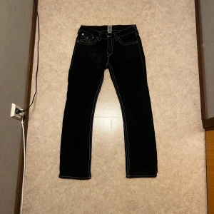True Religion svarta straight jeans 32 - Svarta jeans från True Religion med raka ben och kontrasterande vita sömmar. Klassiska bakfickor med ikonisk brodyr och logopatch bak i midjan. Tillverkade i USA och i bomullsmaterial. Perfekta för dig som gillar streetwear och vill sticka ut.