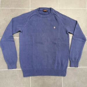 Blå stickad tröja från Morris - Snygg blå stickad tröja från Morris med rund halsringning och diskret broderad logga på bröstet. Tröjan har ribbade muddar vid ärmslut och nederkant, perfekt för lager-på-lager under kyliga dagar.