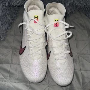 Nike vita fotbollsskor med regnbågssula - Säljer ett par sjukt snygga vita Nike fotbollsskor med regnbågsfärgad sula och svart/rosa swoosh. Skorna har textdetaljer på sidan och coolt rutmönster på ovandelen. Högre strumpliknande skaft och snörning för bästa passform. Perfekta för dig som vill sticka ut på planen!