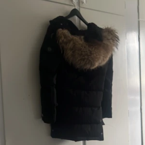Svart dunjacka med päls från Hollies - Säljer en svart dunjacka från Hollies med stor fluffig fuskpäls på huvan. Jackan är quiltad och har en klassisk vinterlook med snygga detaljer och logga på ärmen. Perfekt för kalla dagar när du vill vara både varm och stilren. ❄️