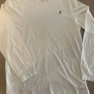 Vit långärmad tröja från Polo Ralph Lauren - Vit långärmad tröja från Polo Ralph Lauren i mjuk bomull. Klassisk design med rund hals och den ikoniska broderade loggan på bröstet. Perfekt basic att ha i garderoben och lätt att matcha med allt.