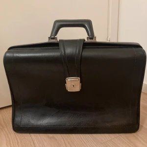 Portfölj i äkta läder - 159 kr  - Klassisk svart portfölj i skinn med robust handtag och silverfärgat lås framtill. Stilren och tidlös design, perfekt för att bära dokument eller laptop. Väskan har en hård struktur och tydliga sömmar längs kanterna för extra stabilitet.