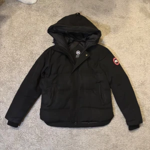 Canada goose Macmillan jacka  - Säljer en svart dunjacka från Canada Goose med huva och klassisk logga på ärmen. Jackan har dragkedja framtill, två fickor och ribbade muddar. Perfekt för kalla dagar och riktigt snygg jacka. Endast använd en vinter och är i riktigt bra skick, vid funderingar är det att höra av sig privat!