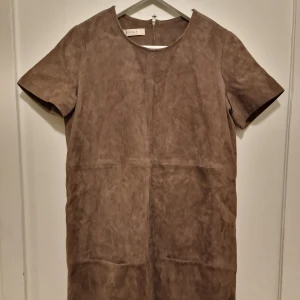 Brun mockaklänning från Sessùn - Snygg brun kortklänning i mocka från Sessùn med rund halsringning, korta ärmar och synliga sömmar samt 2 stora fyrkantig fickor fram. Snygg i kapselgarderoben för en stilren och tidlös design.  Liten M och passar även mig som har S beroende på hur man vill att den ska sitta. 