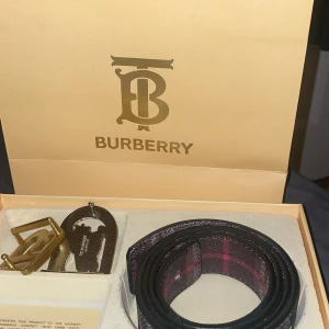 Rutig Burberry bälte med silverdetaljer - Säljer ett snyggt Burberry bälte i klassiskt rutigt mönster med svarta, gråa och röda toner. Bältet har en exklusiv silverfärgad spänne med Burberrys ikoniska riddare. Tillverkat i skinn och levereras i originalförpackning. Perfekt accessoar för att lyfta din outfit.