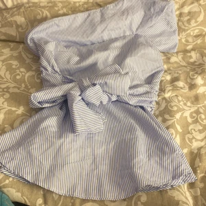 Blåvit randig blus - Supersöt kort blus i blåvitt randigt med knyt i midjan och offshoulder-modell med en axel. Perfekt för varma dagar och har en luftig känsla. Gjord i mjuk bomull som känns skön mot huden. Snygg och trendig look med elastisk detalj för extra komfort. Aldrig använt denna, men den har ett hål vid dragkedjan men kan enkelt sys ihop💓 