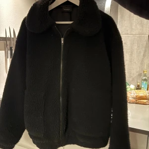 Svart teddyjacka med dragkedja - Mysig svart teddyjacka från tindra med stor krage och dragkedja framtill. Jackan har två stora fickor och en loose passform som ger en chill vibe. Perfekt för kalla dagar när du vill vara både varm och snygg.