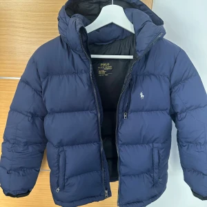 Mörkblå pufferjacka från Polo Ralph Lauren - Säljer en mörkblå pufferjacka från Polo Ralph Lauren med huva och dragkedja. Jackan har klassisk quiltad design, två sidofickor med dragkedja och Polo-logga broderad på bröstet. Perfekt för kalla dagar och riktigt snygg till streetwear.