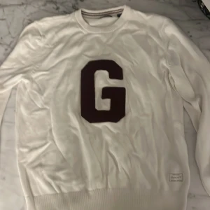 Vit tröja med GANT-logga och stor G - Vit långärmad tröja från GANT med en stor mörkröd bokstav G på bröstet. Tröjan har ribbade muddar vid ärmslut och nederkant samt en liten GANT-logga nertill. Perfekt för dig som gillar klassisk och stilren design.