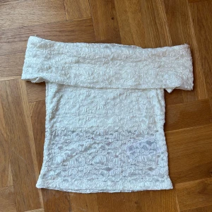 Vit offshoulder spets tubtopp - Säljer en vit offshoulder tubtopp i spets med blommigt mönster. Toppen är croppad och har en bred kant upptill som sitter snyggt över axlarna. Perfekt att styla med jeans eller kjol för en trendig look.