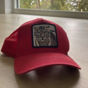 Röd keps Goorin Bros Tiger - Röd keps från Goorin Bros. med mesh på sidorna och bak, samt broderad tigerpatch framtill. Justerbar passform och klassisk böjd skärm. Märket och designen ger en cool och sportig vibe, perfekt för att sticka ut i mängden.