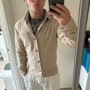 Beige överskjortq från Ciszere - Snygg beige skjorta från Ciszere. Skjortan har klassisk krage, bröstficka och knäppning framtill med träfärgade knappar. Slim fit-modell med långa ärmar, perfekt för en clean och avslappnad look. Bra skick 