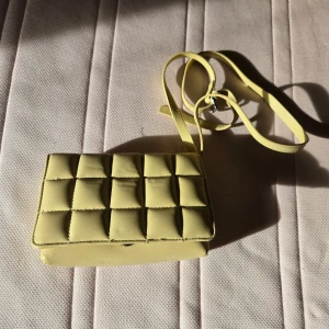 Gul quiltad axelväska i skinnimitation - Trendig gul axelväska med quiltad design och lång axelrem. Väskan har en stilren, rektangulär form och är tillverkad i skinnimitation. Perfekt för att lyfta din outfit med en poppig färg och cool struktur.