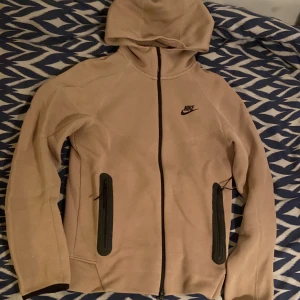 Beige Nike tech fleece, XS - Säljer en beige hoodie från Nike i storlek XS. Tröjan har hel dragkedja, två svarta fickor med dragkedja och klassisk Nike-logga på bröstet. Utrustad med huva och långa ärmar, perfekt för chill eller träning. Nästan aldrig använd, kan förekomma katthår tyvärr annars helt felfri.