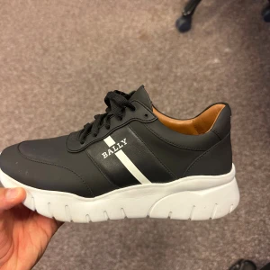 Svarta Bally sneakers med vit sula - Snygga svarta sneakers från Bally med chunky vit platt sula och svarta snören. Skorna har en vit rand och Bally-logga på sidan samt bak på hälen. Utsidan är i skinn och insidan har en brun läderdetalj. Perfekta för dig som gillar stilrena och sportiga skor. Använda 1 gång. Nypris 7500:-  Material Yttre: kalvskinn 82%, lammskinn 18%  Sula: gummi 100%  Foder: Tyg 100%  https://www.farfetch.com/se/shopping/men/bally-sneakers-med-banddetalj-item-30411327.aspx