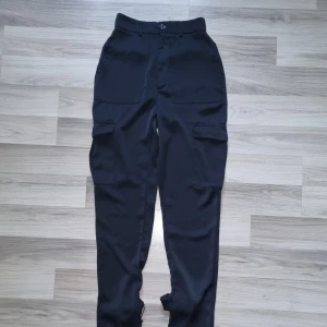 Svarta cargopants från BikBok, strl 34 - Svarta cargopants från BikBok med flera stora fickor på sidorna och bak, samt snygga spänndetaljer vid bensluten. Byxorna har hög midja och är tillverkade i ett mjukt, lätt material som ger en chill och trendig look.