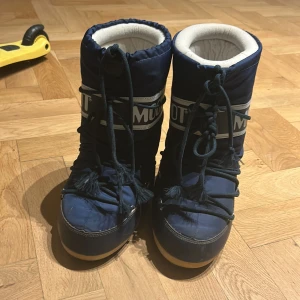 Blå Moon Boots med snörning - Säljer ett par klassiska blå Moon Boots med vit insida och tydlig logga runt skaftet. Skorna har snörning framtill och är tillverkade i syntetmaterial med en platt sula. Perfekta för vintern och håller fötterna varma och torra. OBS En av skorna är sönder där bak. Se  sista bilden .