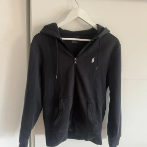 Svart hoodie från Ralph Lauren - Svart hoodie från Ralph Lauren med dragkedja framtill, huva med snörning och klassisk vit logga på bröstet. Mjuk bomullskvalitet och två fickor framtill.