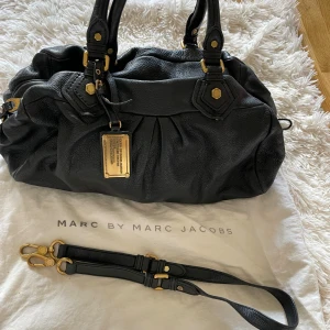 Svart skinnväska Marc by Marc Jacobs - Snygg svart handväska från Marc by Marc Jacobs i äkta skinn med guldfärgade metalldetaljer och avtagbar axelrem. Väskan har plisserade detaljer, dekorativ metallbricka framtill och innerfoder med logomönster. Perfekt för dig som vill ha en klassisk och stilren väska.