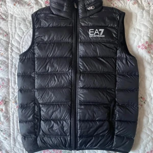 Svart dunväst från EA7 Emporio Armani  - Snygg svart dunväst från EA7 Emporio Armani med quiltad design och logga på bröstet. Västen har dragkedja framtill och avtagbar huva. Perfekt för lager-på-lager och streetstyle.
