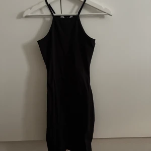 Svart figurnära klänning  - En svart, tight och kort klänning från Nelly med smala axelband och hög halsringning. Klänningen har en minimalistisk design och är perfekt för dig som gillar en stilren look🖤