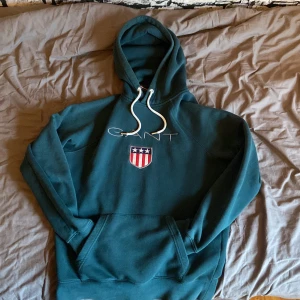Mörkgrön hoodie från GANT med brodyr - Säljer en mörkgrön hoodie från GANT med broderad logga och sköld på bröstet. Tröjan har huva med vita snören och en stor magficka. Insidan av huvan har en snygg randig detalj i rött, vitt och blått. Perfekt för chill dagar.